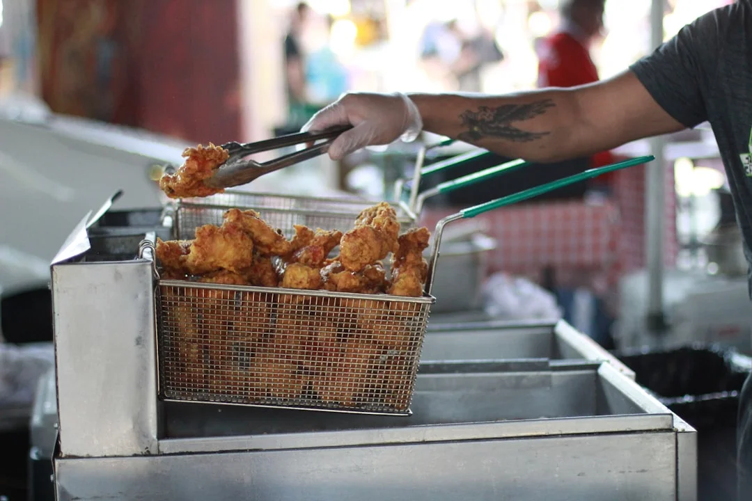 Close-up of a heavy-gauge stainless steel twin fryer basket set resting on the edge of a commercial countertop deep fryer, golden-fried food visible inside, professional kitchen tiles in the background
